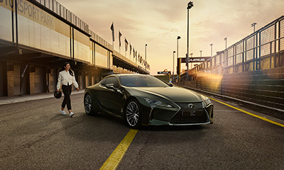A woman dressed in a white jacket and black pants and holding a helmet walks towards an LC coupe parked in a pit lane.