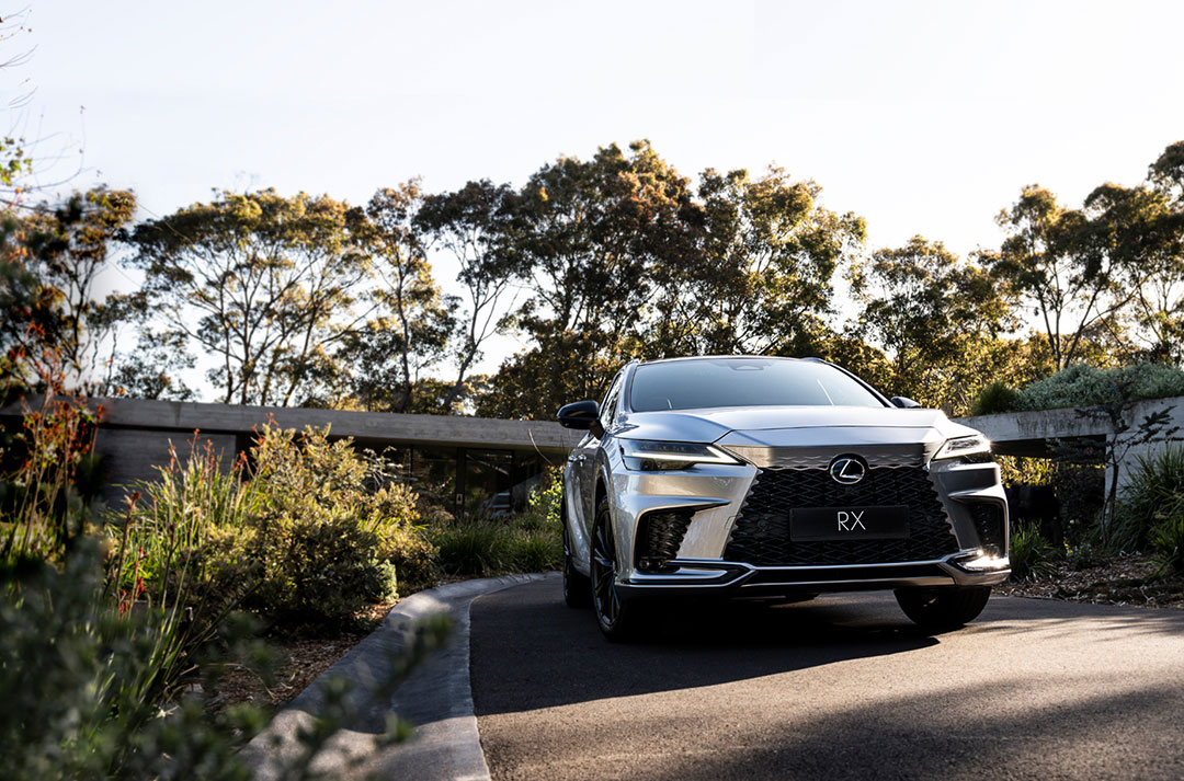 A silver Lexus RX 500h is parked on a driveway, native Australian bushes and blue sky are around it.