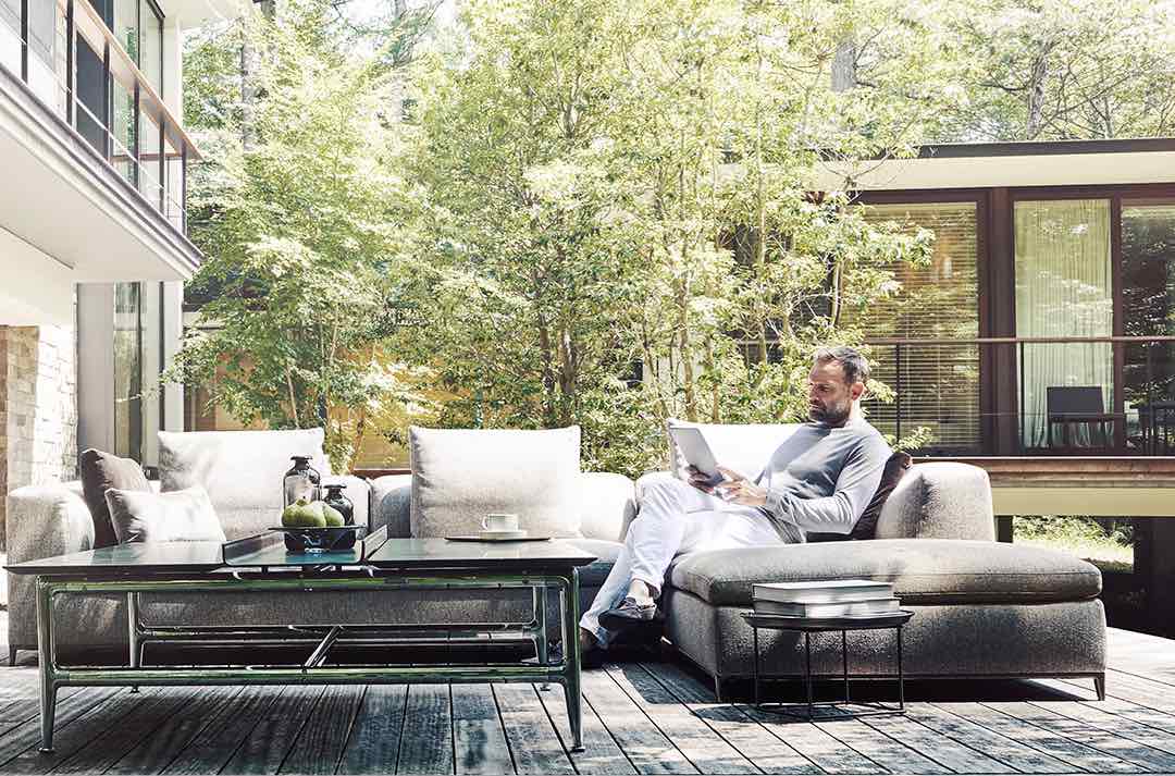 Man sitting on a couch in his backyard, reading a paper. Greenery surrounds him.