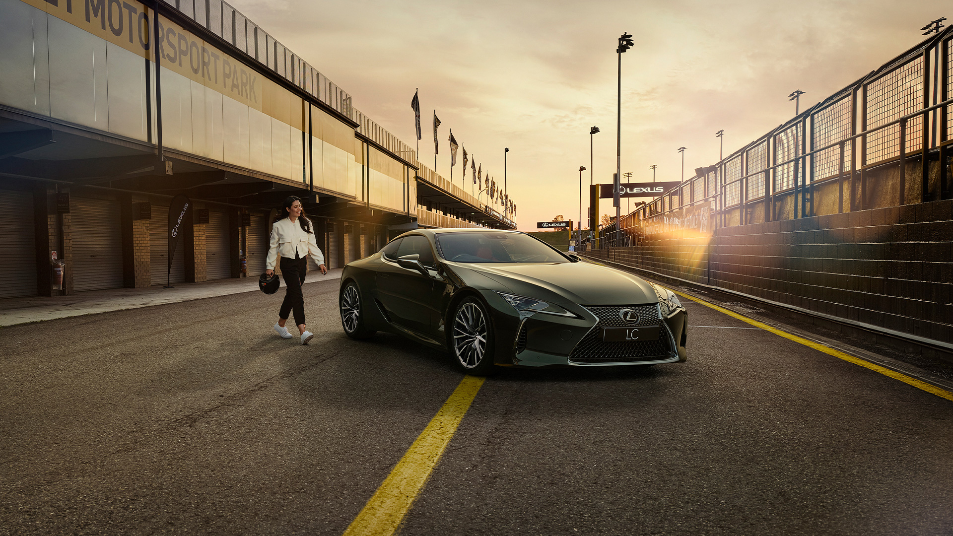 A woman dressed in a white jacket and black pants and holding a helmet walks towards an LC coupe parked in a pit lane