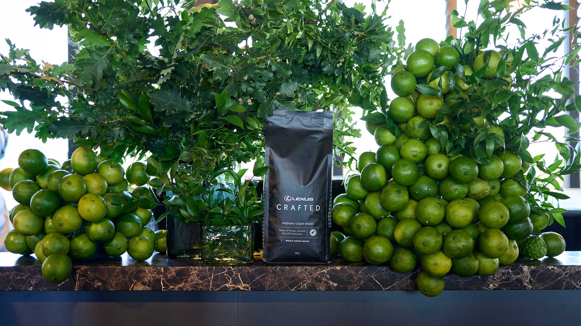 A bag of Lexus Crafted coffee beans displayed between arrangements of green citrus fruit and foliage.