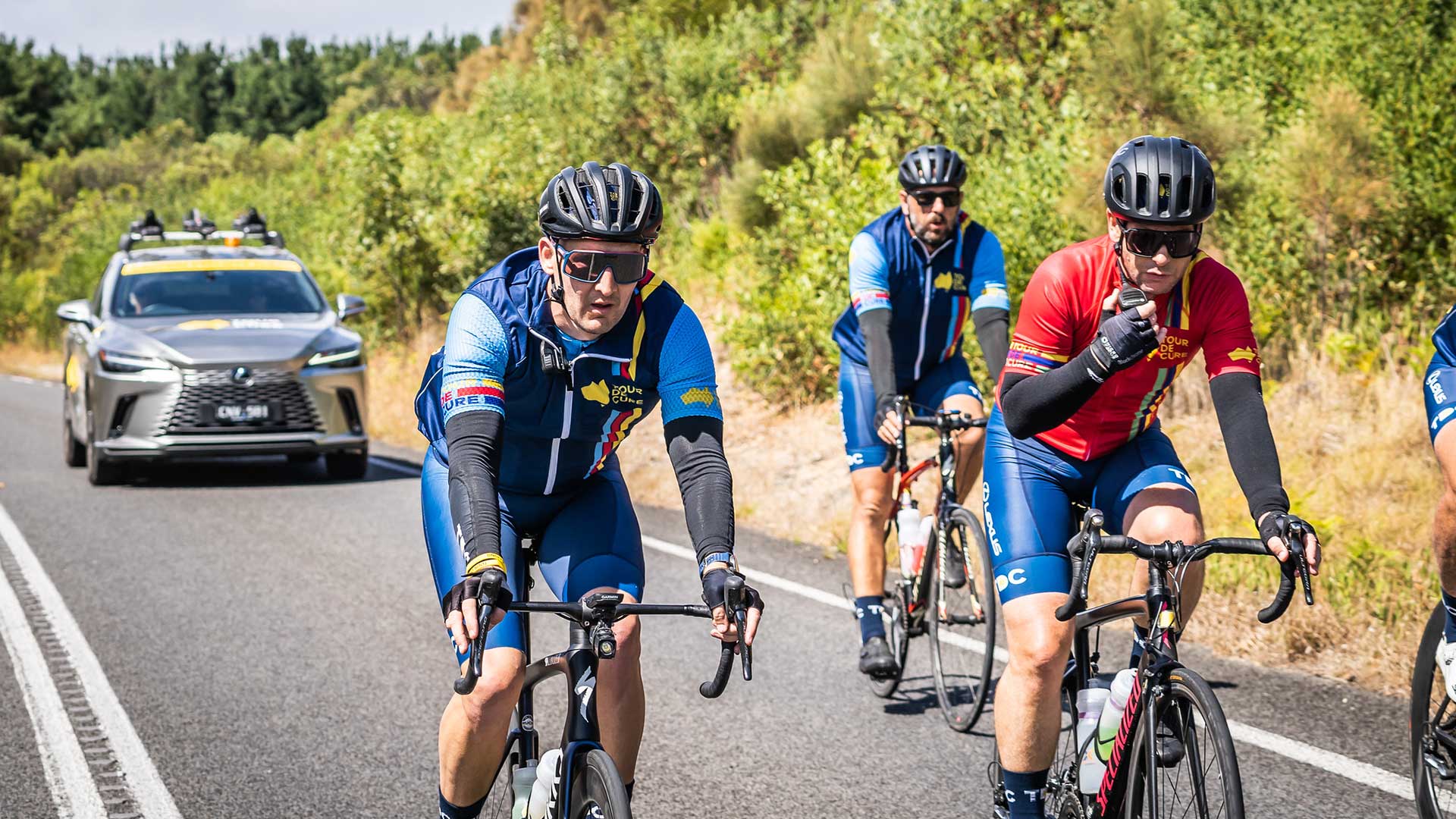 A group of cyclists ride together along a rural road, with Lexus support vehicle following behind.