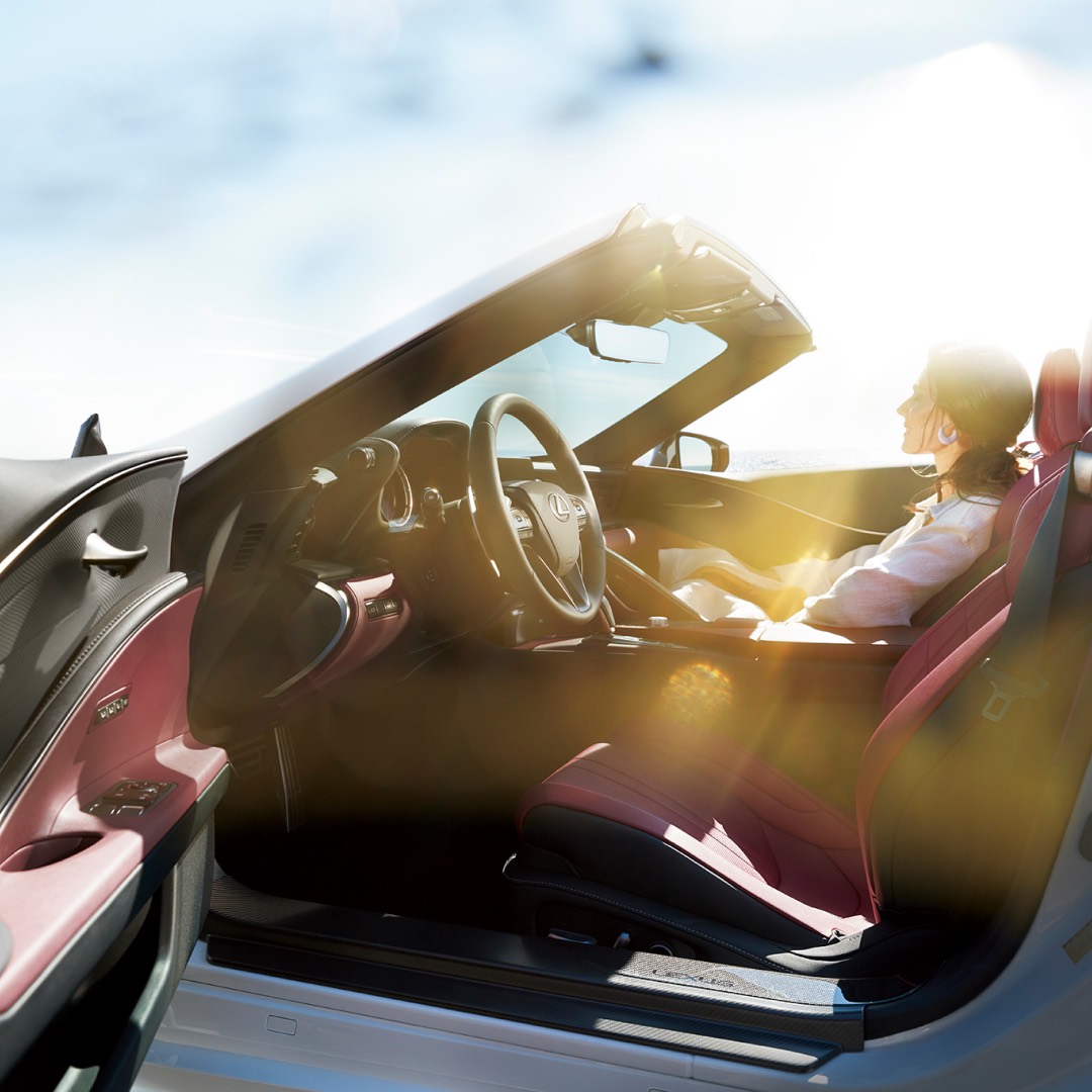 Interior view of a Lexus LC Convertible with red leather seats, a woman in the passenger seat with sunlight streaming in.