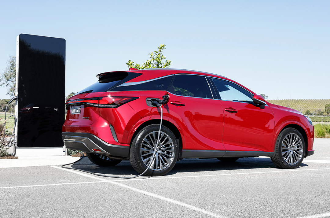Red Lexus RX 450h+ plug-in hybrid charging at an electric vehicle station in an outdoor setting.
