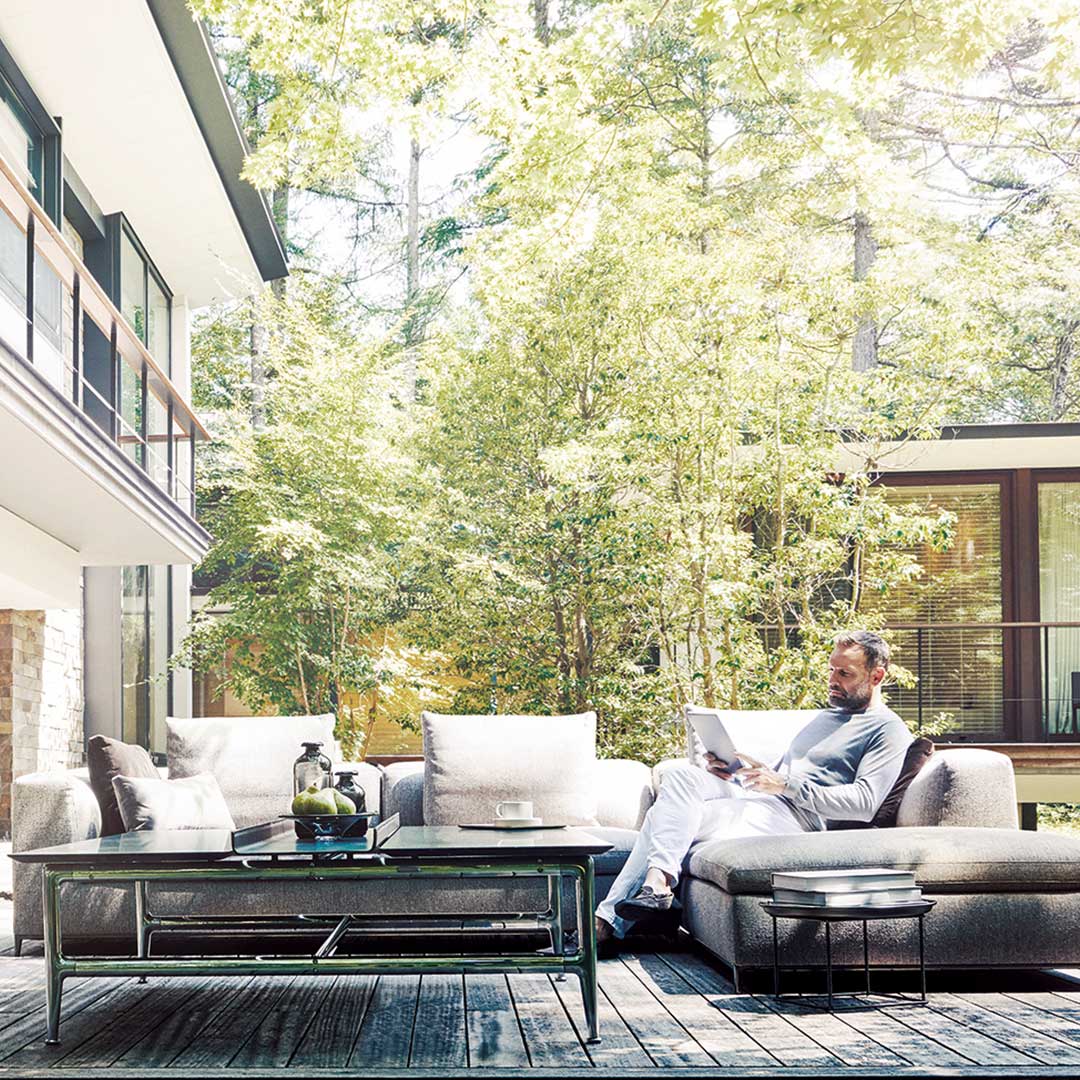 Man sitting on a couch in his backyard, reading a paper. Greenery surrounds him.